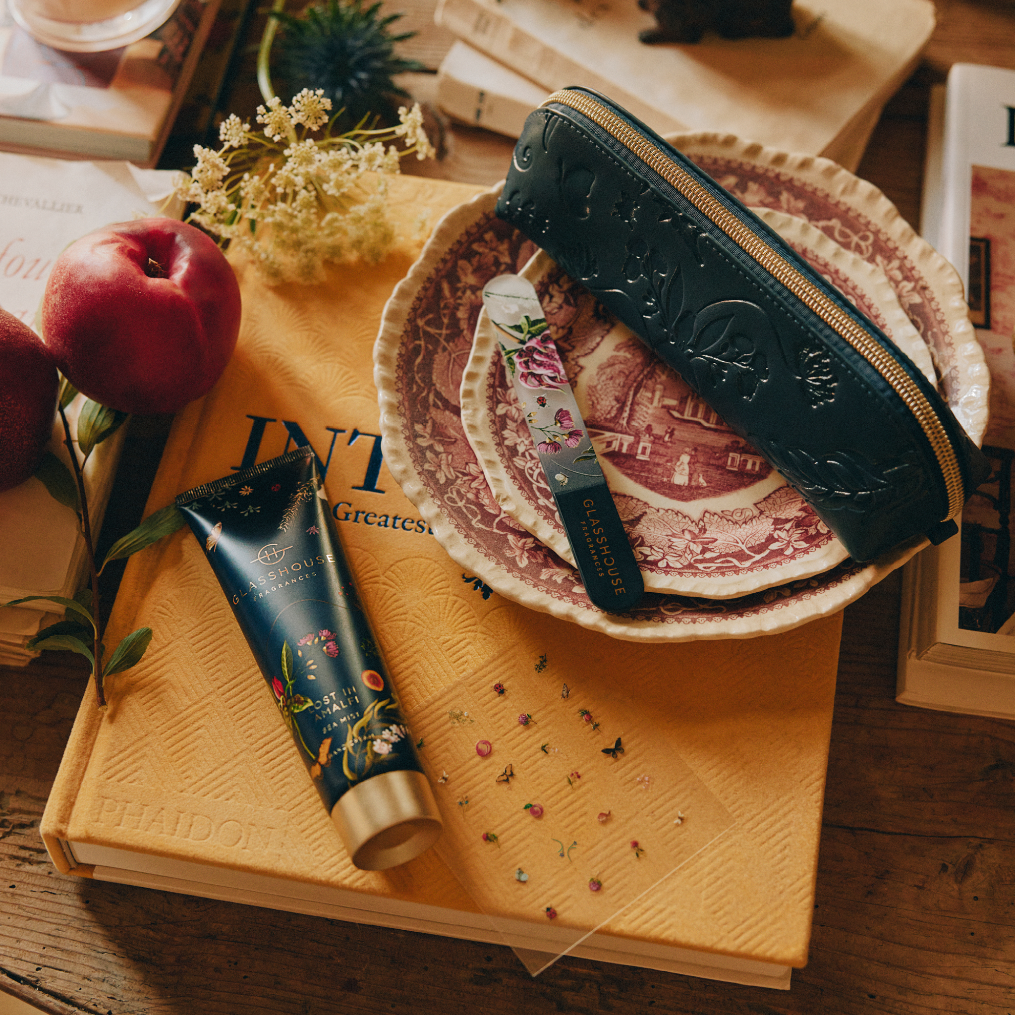 Glasshouse Fragrances Lost In Amalfi Hand & Nail Set, blue cosmetic pouch, Hand Cream, Glass Nail File and decorative Nail Stickers on table with book and fruit.