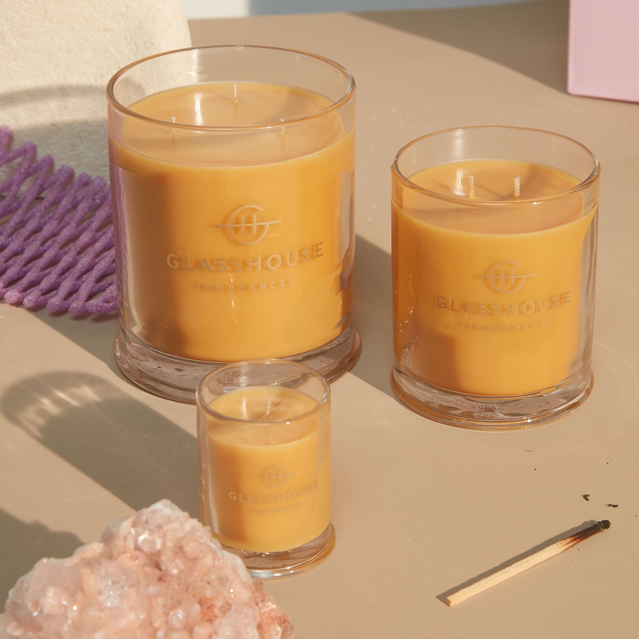 Three various sizes of A Tahaa Affair Vanilla Caramel Soy Candle burning on a tabletop beside a crystal